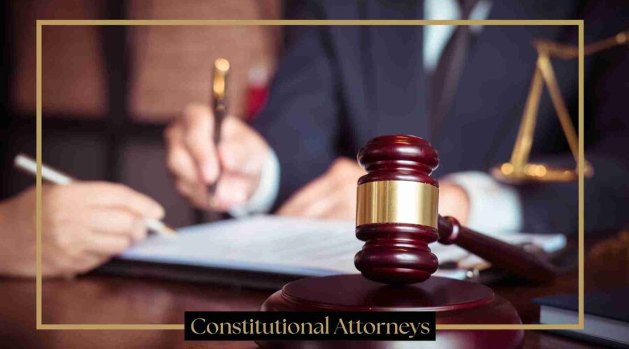 A judge's gavel on a wooden desk in a courtroom setting, with an attorney writing in the background and the text "Constitutional Attorneys" at the bottom.