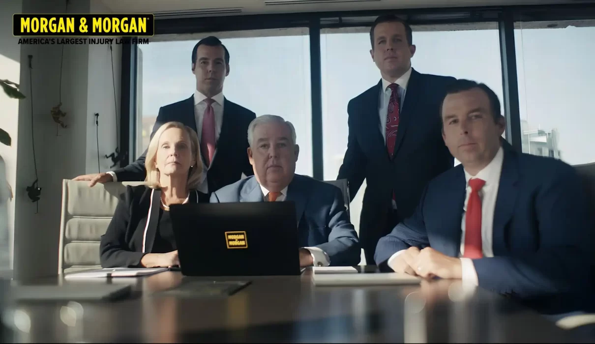 A group portrait of five attorneys from the Morgan & Morgan law firm, including founder John Morgan, seated and standing around a conference table in a modern office.