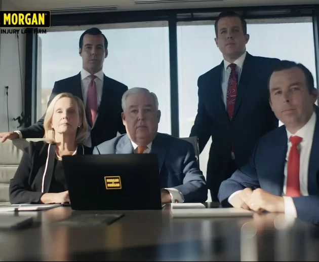 A group portrait of five attorneys from the Morgan & Morgan law firm, including founder John Morgan, seated and standing around a conference table in a modern office.