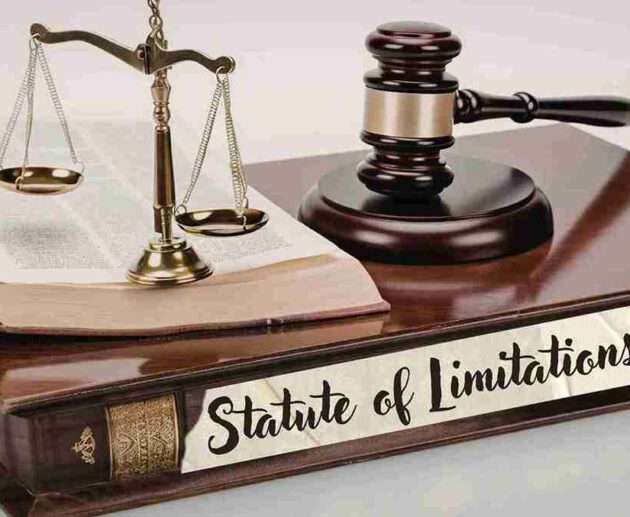 A legal-themed still life featuring the Scales of Justice, a judge's gavel, and an open law book resting on a large, leather-bound volume. A white paper label on the front of the book features the handwritten text "Statute of Limitations"