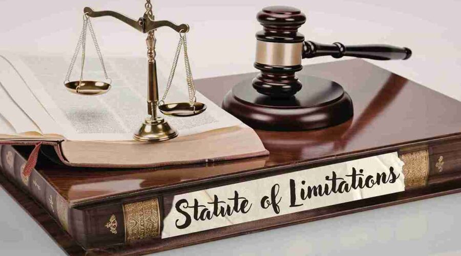 A legal-themed still life featuring the Scales of Justice, a judge's gavel, and an open law book resting on a large, leather-bound volume. A white paper label on the front of the book features the handwritten text "Statute of Limitations"