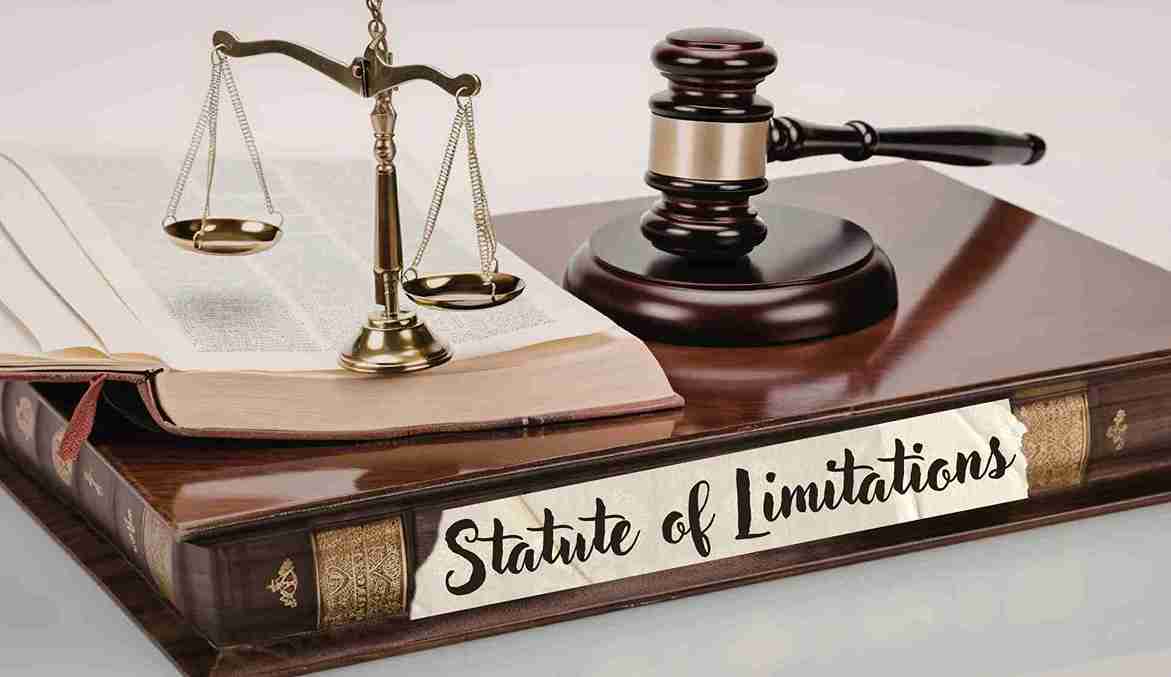 A legal-themed still life featuring the Scales of Justice, a judge's gavel, and an open law book resting on a large, leather-bound volume. A white paper label on the front of the book features the handwritten text "Statute of Limitations"