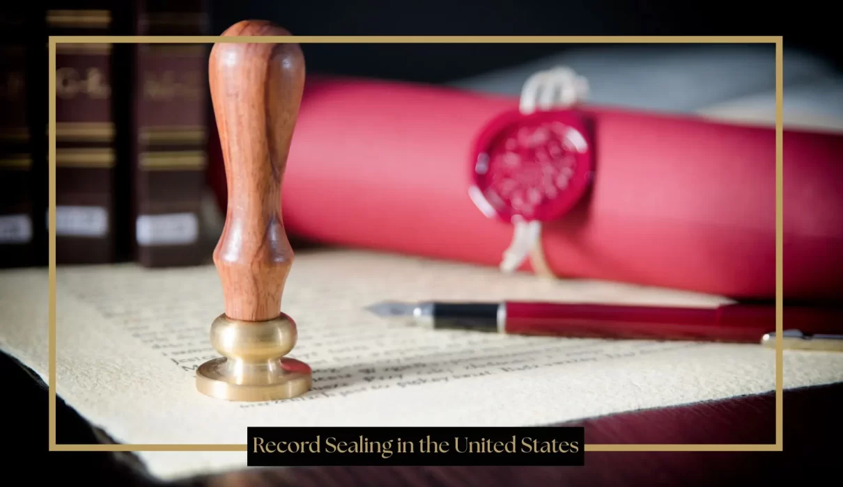 A wooden and brass wax seal stamp stands upright on a piece of parchment paper next to a fountain pen and a rolled red document secured with a red wax seal.