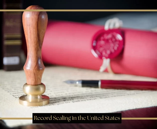 A wooden and brass wax seal stamp stands upright on a piece of parchment paper next to a fountain pen and a rolled red document secured with a red wax seal.