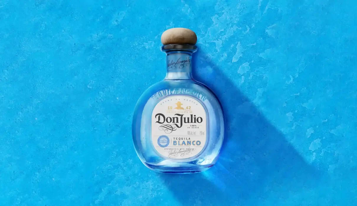 A bottle of Don Julio Blanco Tequila—subject of false advertising class-action lawsuits over its "100% Agave" labeling—centered against a textured blue background. The rounded glass bottle features a natural wood cork stopper and a white front label showing the prominent branding. A soft shadow drops diagonally to the lower right.