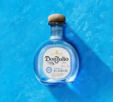 A bottle of Don Julio Blanco Tequila—subject of false advertising class-action lawsuits over its "100% Agave" labeling—centered against a textured blue background. The rounded glass bottle features a natural wood cork stopper and a white front label showing the prominent branding. A soft shadow drops diagonally to the lower right.