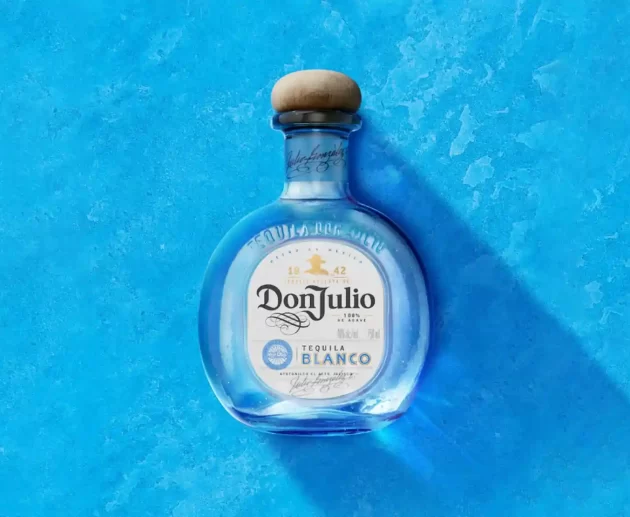 A bottle of Don Julio Blanco Tequila—subject of false advertising class-action lawsuits over its "100% Agave" labeling—centered against a textured blue background. The rounded glass bottle features a natural wood cork stopper and a white front label showing the prominent branding. A soft shadow drops diagonally to the lower right.