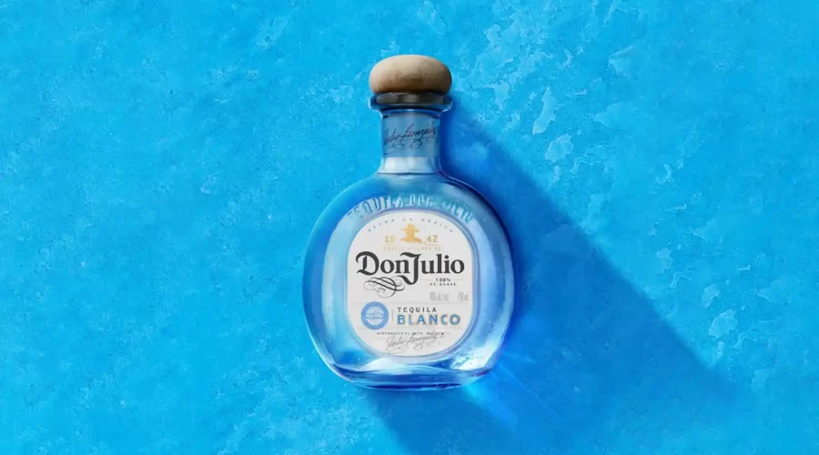 A bottle of Don Julio Blanco Tequila—subject of false advertising class-action lawsuits over its "100% Agave" labeling—centered against a textured blue background. The rounded glass bottle features a natural wood cork stopper and a white front label showing the prominent branding. A soft shadow drops diagonally to the lower right.